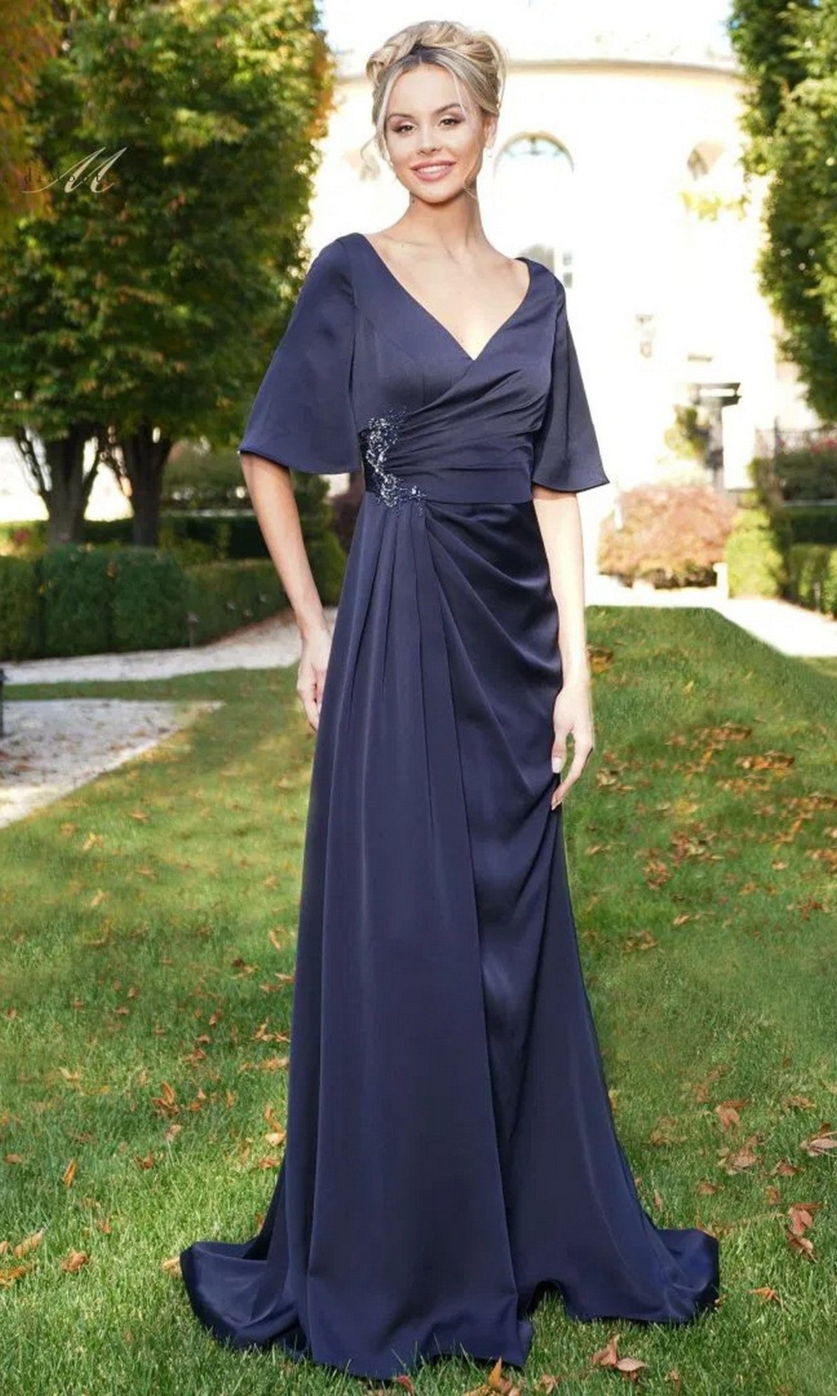 A woman poses outdoors on grass in front of a building and trees, wearing the navy Rina Di Montella RD2974 long formal dress featuring a ruched fitted bodice and crepe floor-length design.